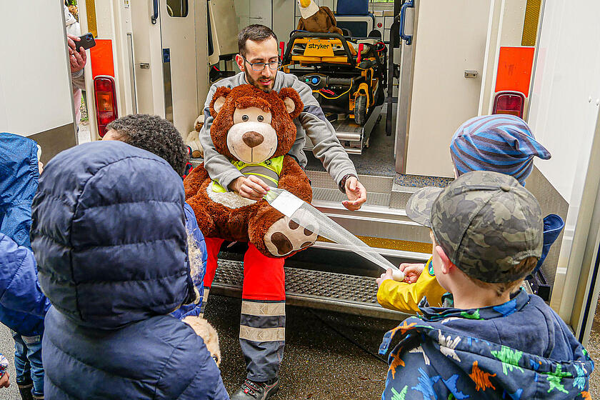 Ein Johanniter hat einen großen Teddybären auf dem Schoß und sitzt hinten im Eingang eines Rettungswagens. Ihm schauen mehrere Kinder zu, eines davon hält ihm eine Rolle Verband hin, welchen er gerade abrollt.