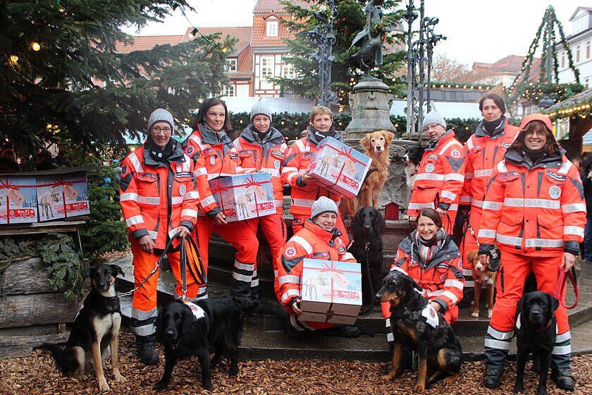 Mitglieder und Hunde der Rettungshundestaffel Südniedersachsen zeigen ihre gepackten Pakete. Im Hintergrund sind die Tannen und Lichterketten des Weihnachrtsmarktes zu sehen.