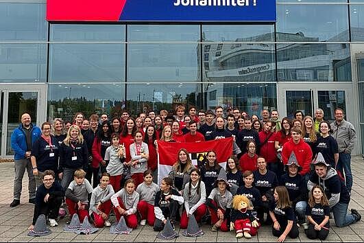 Gruppe von Johanniter-Mitgliedern, bestehend aus Kindern, Jugendlichen und Erwachsenen, posiert lächelnd vor der Messe in Erfurt. Im Hintergrund ist auf einem großen digitalen Display das Johanniter-Logo zu sehen, dazu die Botschaft: „Wer sind die besten Rettungskräfte im ganzen Land? – Na klar, wir Johanniter!“. In der ersten Reihe halten mehrere Teilnehmende eine rot-weiße Flagge, vorne sitzt ein Maskottchen in Johanniter-Kleidung.