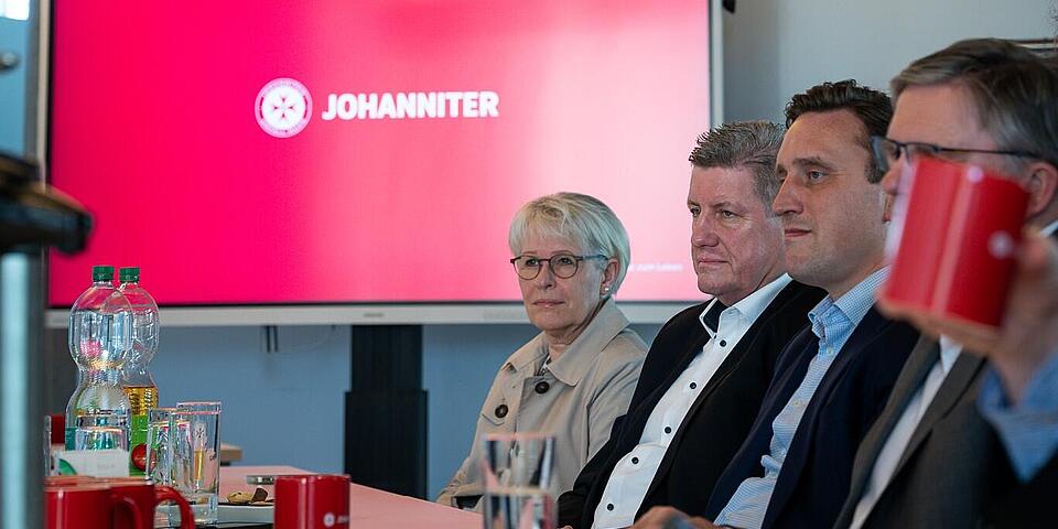 Menschen an einem Konferenztisch. Im Hintergrund ein großer Bildschirm auf dem der Schriftzug Johanniter mit roter Hintergrundfarbe zu sehen ist