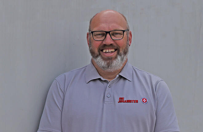 Ein Mann mit Halbglatze und graumeliertem Vollbart und Brille. Er trägt ein graues Poloshirt mit Johanniter-Logo