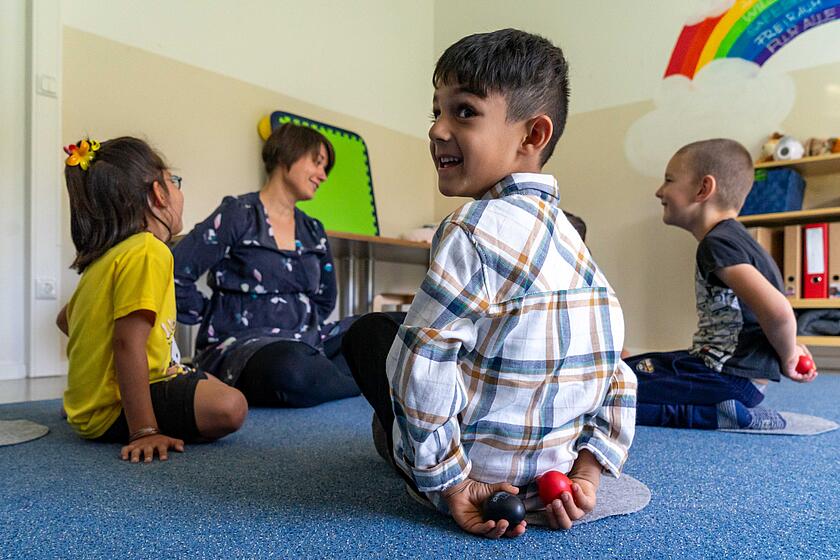 Ein Junge hat Klangeier in der Hand, die er hinter dem Rücken versteckt. Er sitzt mit anderen Kindern und einer Musikpädagogin auf dem Boden in einem Kreis.