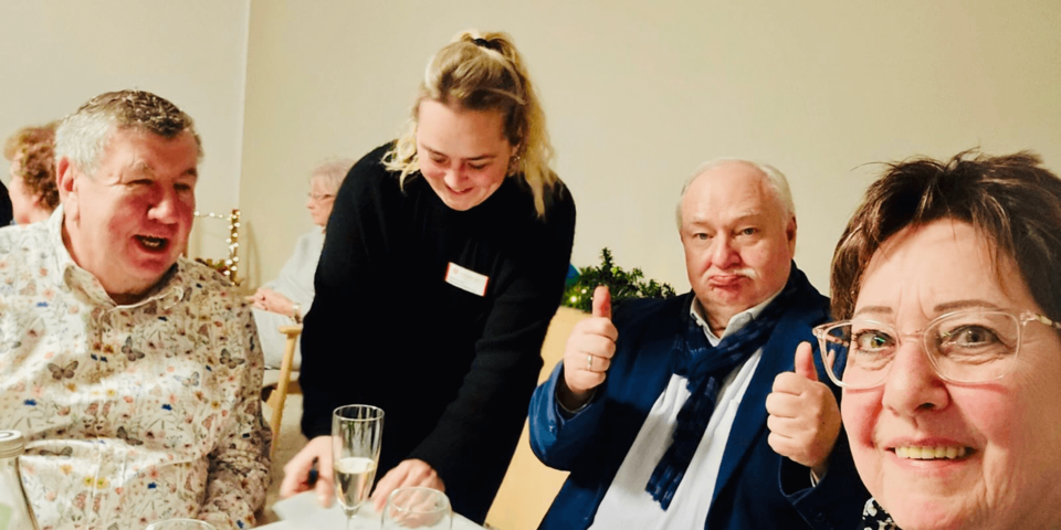 Gäste an einem festlich gedeckten Tisch, eine Servicekraft serviert, ein Gast zeigt Daumen nach oben, fröhliche Stimmung.
