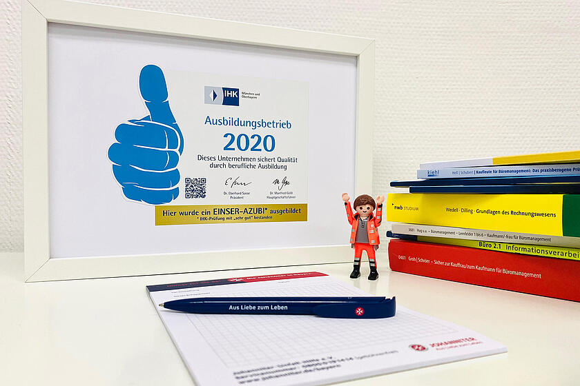 Ein eingerahmtes Zertifikat der IHK für die Johanniter-Unfall-Hilfe für einen Einser-Schüler steht neben einem Block, einem Stift, einem Johanniter Playmobilmännchen und einem Stapel an Lernbüchern auf einem Tisch.