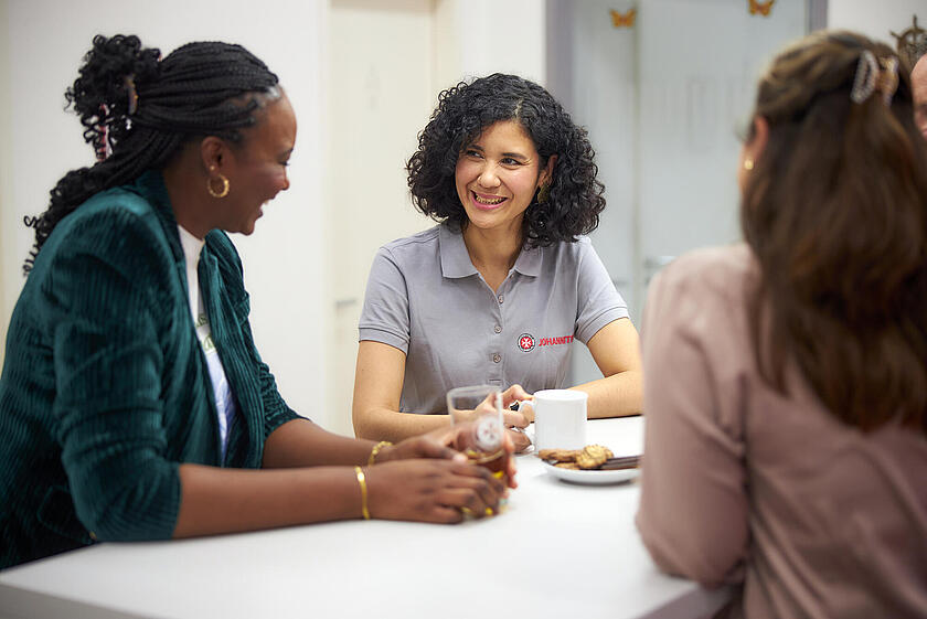 3 Frauen sitzen an einem Tisch mit Getränken und Keksen und lächeln.