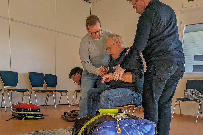 Zwei Männer kümmern sich um einen älteren Mann, der auf einem Stuhl sitzt und Probleme mit der Atmung hat. Ein weiterer Mann kniet über einem Rucksack.