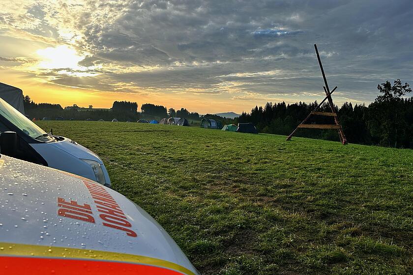 Auf einer großen Grünfläche ist ein Zeltlager aufgebaut. Links stehen Johanniter Einsatzfahrzeuge und am Himmel scheint die Sonne zwischen den Wolken hervor.