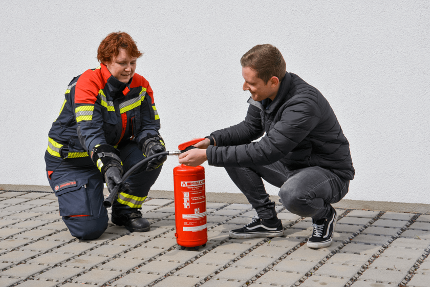 Ein Frau in Einsatzkleidung und ein Mann in dunkler Privatkleidung knieen auf Setinplatten neben einem Feuerlösche rund sprechen darüber. 