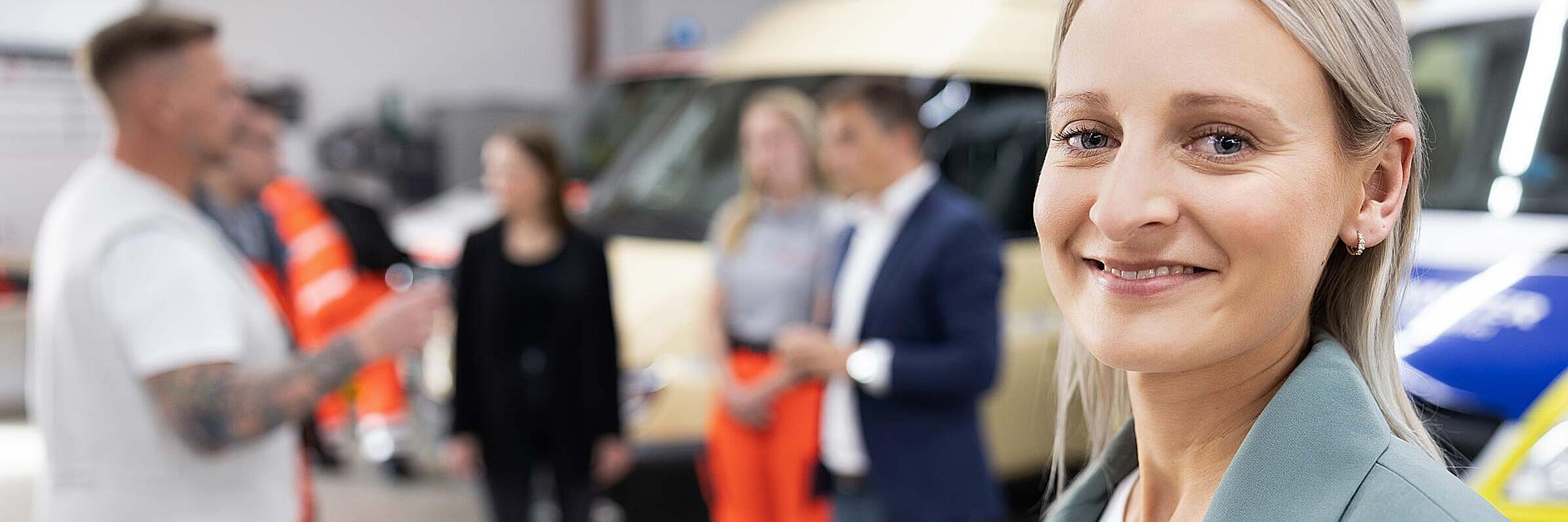 Frau im grünen Blazer steht vor Einsatzfahrzeugen, im Hintergrund mehrere Personen im Gespräch. 
