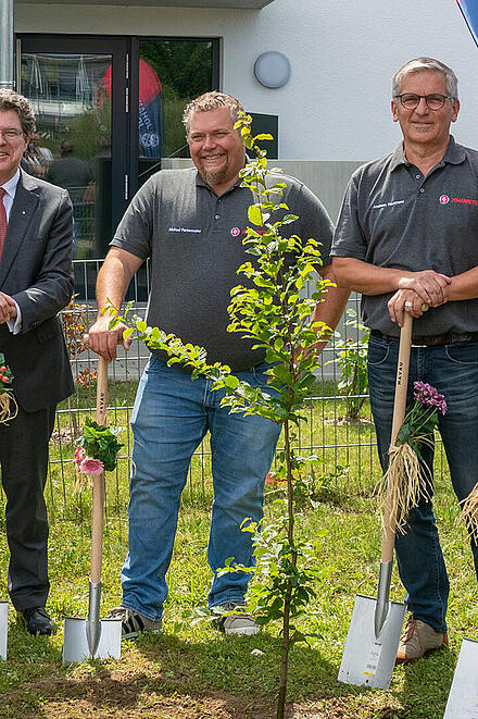 Ein Johanniter-Pfarrer und 5 weitere Johanniter stehen bei einer feierlichen Baumpflanzaktion vor einem Gebäude. Der Pfarrer hält eine Gießkanne in der Hand, die anderen halten jeweils einen geschmückten Spaten. Im Hintergrund ist eine Fahne, auf der "Johanniter" steht.