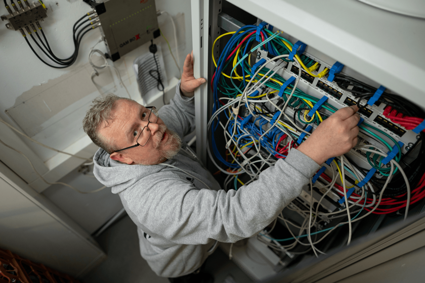 Ein Mann in grauer Arbeitskleidung arbeitet an einem Netzwerkschrank mit bunten Kabeln