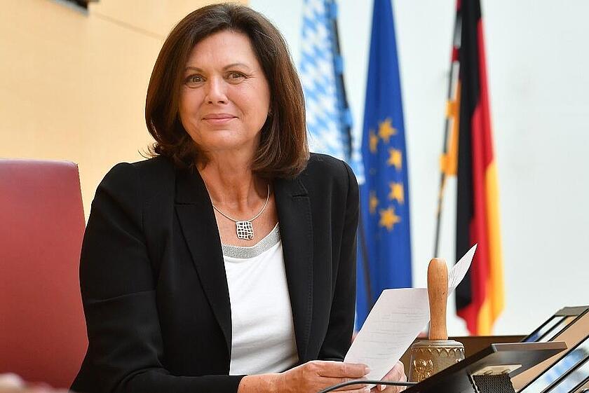 Frau sitzt mit Dokumenten am Rednerpult, hinter ihr Flaggen von Deutschland, Bayern und der EU.