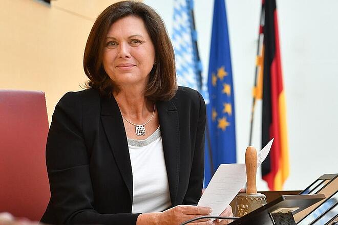 Ilse Aigner Frau sitzt mit Dokumenten am Rednerpult, hinter ihr Flaggen von Deutschland, Bayern und der EU.