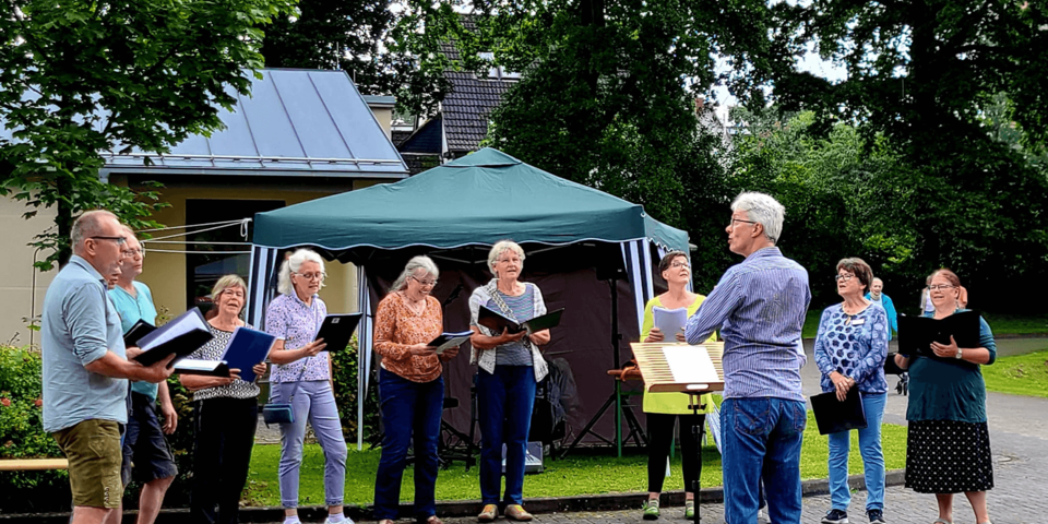 Eine Gruppe steht singend im Freien. Eine Person dirigiert.