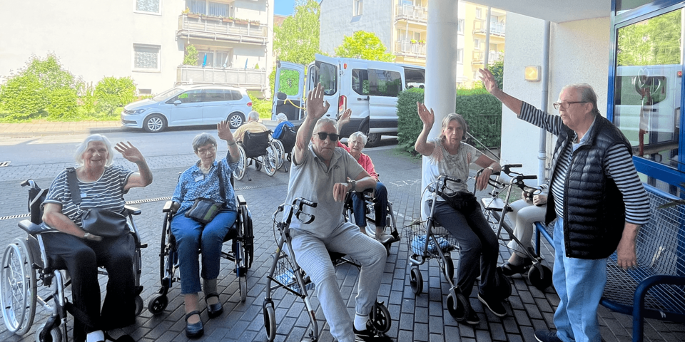 Menschen stehen mit Rollatoren unter einem Vordach und winken