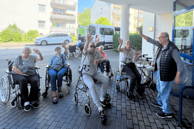 Menschen stehen mit Rollatoren unter einem Vordach und winken