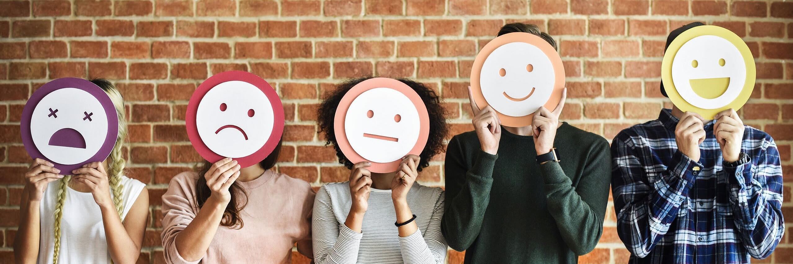 Fünf Leute stehen vor einer Steinmauer und halten bunte Smileys mit verschiedenen Stimmungen vor ihr Gesicht.