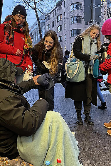 Eine obdachlose Person sitzt mit Decke auf dem Bürgersteig, Helfende verteilen warme Getränke und Schlafsäcke.