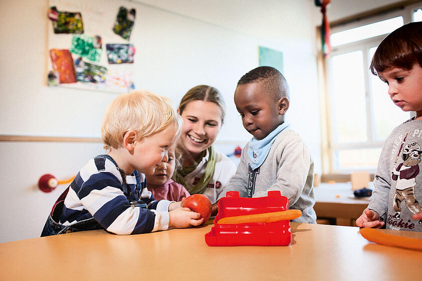 Erzieherin sitzt mit vier Kindern an einem Tisch beim gemeinsamen Basteln und Spielen