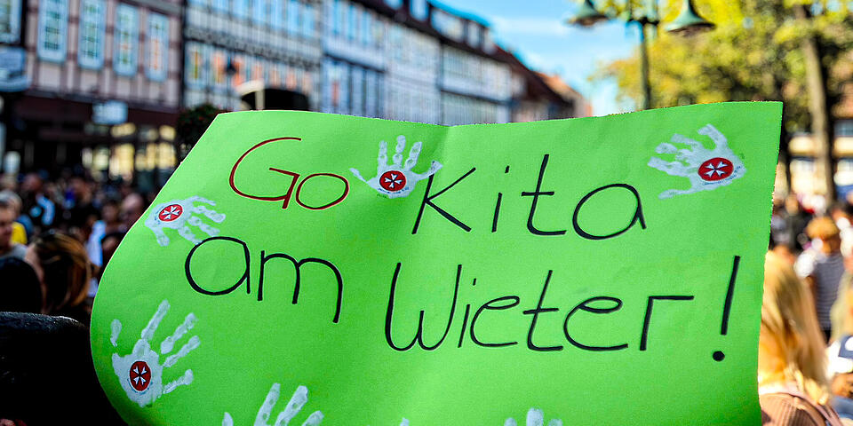 Ein grünes Schild auf dem steht: "Go Kita am Wieter" wird in einer Menschenmenge in einer Innenstadt hochgehalten.