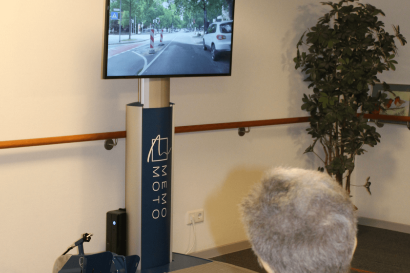 Eine Person ist von hinten dabei zu sehen, wie sie auf einen Bildschirm blickt, auf dem eine Straße zu sehen ist. Links unten im Bild sieht man ein Fußfahrrad.