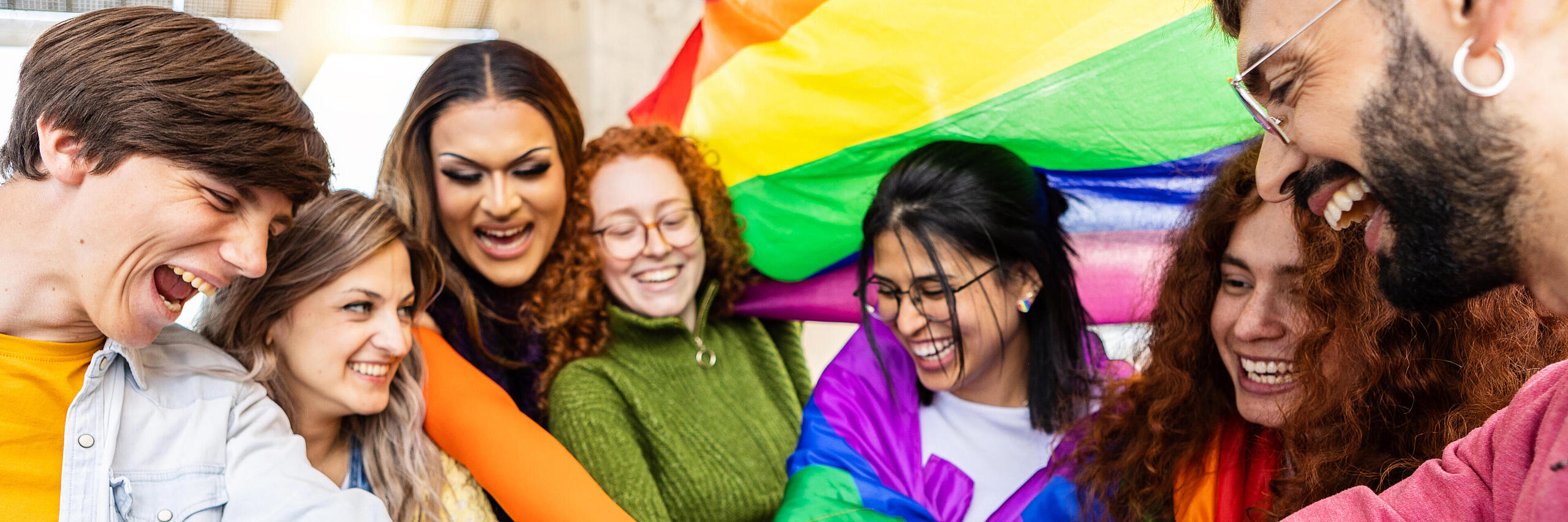 Junge Menschen verschiedenster Identitäten schlagen zusammen im Kreis ein, im Hintergrund weht eine Regenbogenflagge.