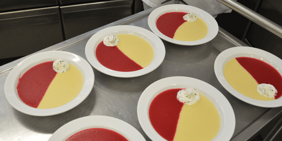 Ein Tablettwagen, auf dem sechs Teller mit Suppe stehen. Die Suppe ist zur Hälfte rot und zur anderen Hälfte hellgelb
