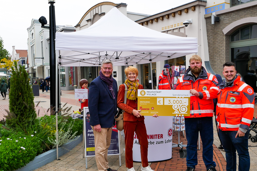 2 Johanniter und zwei andere Leute stehen in Ingolstadt Village und halten einen großen Spendencheck für die Johanniter.