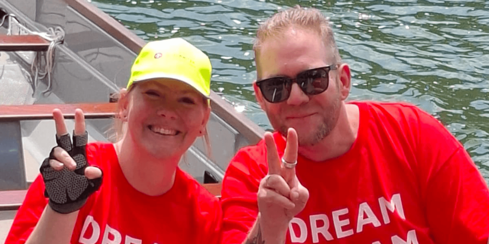 Zwei Personen in roten T‑Shirts zeigen Victory‑Zeichen und lächeln im Boot.