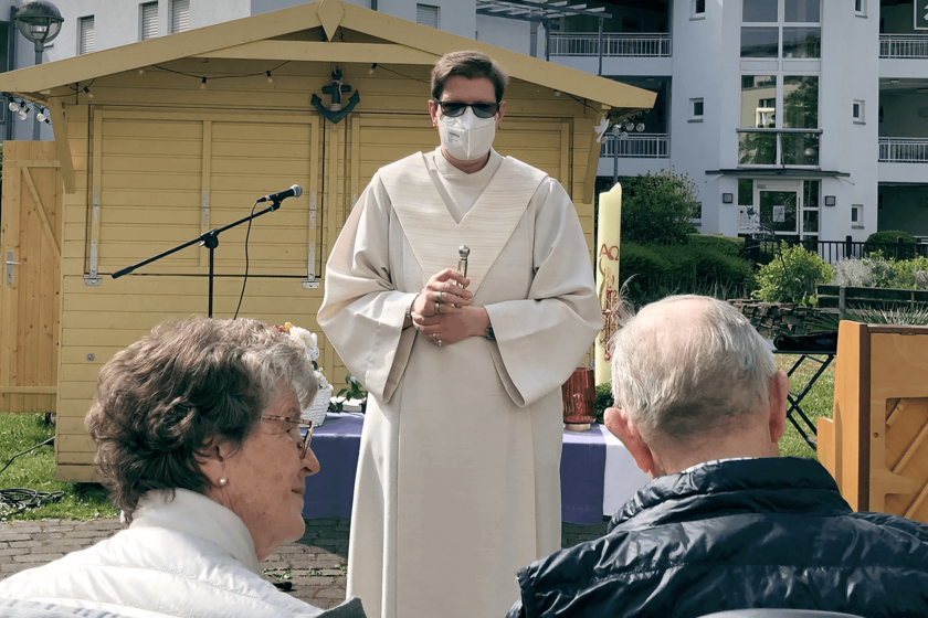 Ein älteres Paar sitzt im Freien einem weißgekleideten Geistlichen gegenüber.