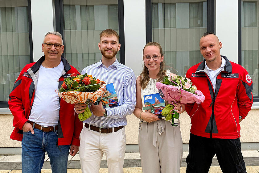 Zwei Männer stehen recht und links. Sie tragen eine Johanniter-Jacke. In der Mitte stehen ein junger Mann und eine junge Frau mit Zertifikaten und Blumensträußen in der Hand