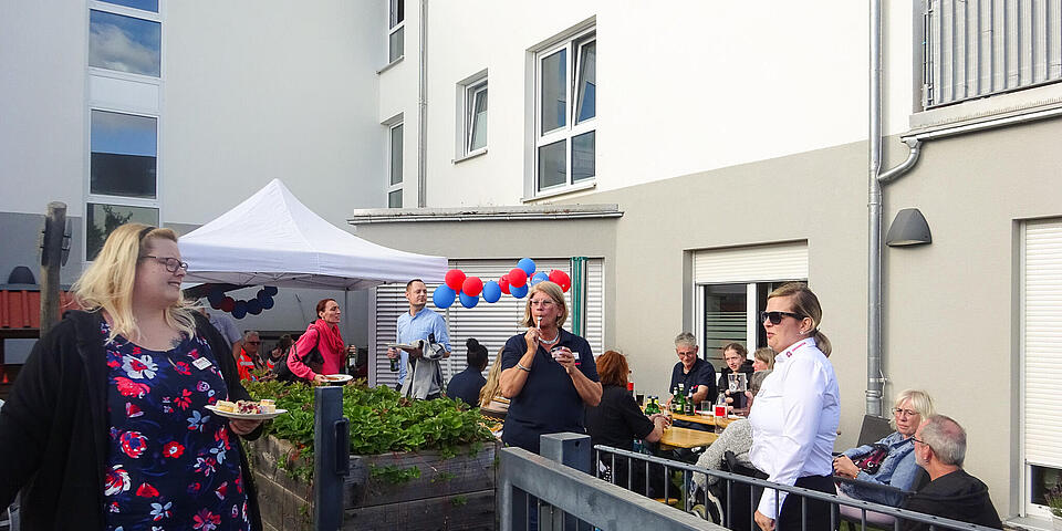 Auf einer Außenanlage vor einem Haus steht ein Pavillon und es sind Menschen auf Bierzeltgarnituren zu sehen.