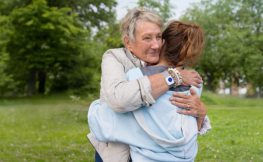 Zwei Frauen umarmen sich. Die ältere Frau trägt einen Handsender am Handgelenk. Im Hintergrund ist ein Park zu sehen.