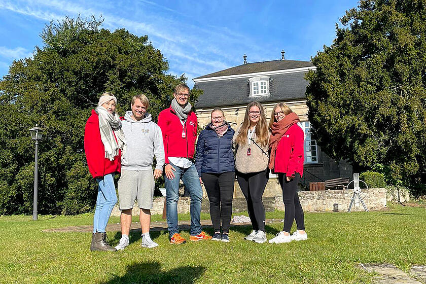 Sechs motivierte junge Erwachsene in Johanniter-Jugend-Kleidung stehen vor einem Landhaus auf einer Wiese und blicken in die Kamera.