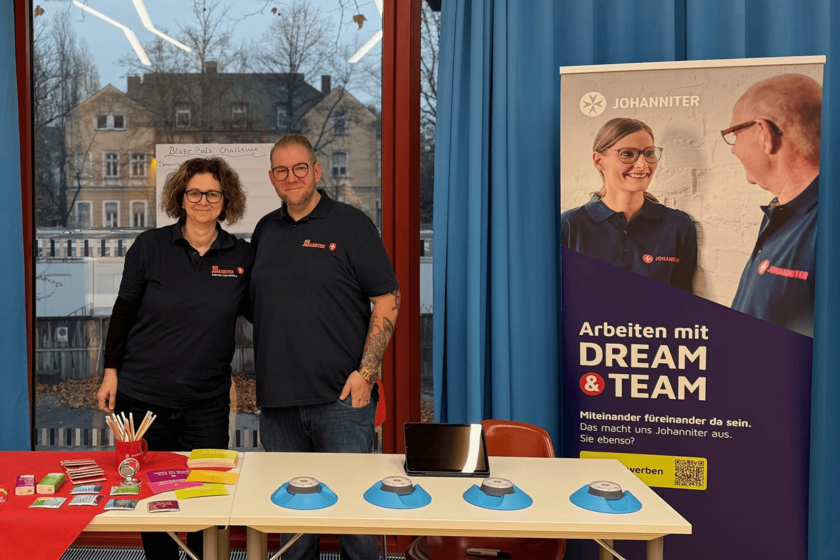Zwei Johanniter-Mitarbeitende stehen zusammen hinter einem Infostand auf einer Messe. Der Stand ist dekoriert mit Flyern, einer roten Johanniter-Tasse und einem weißen Roll-Up mit dem Text "Arbeiten mit DREAM & TEAM".
