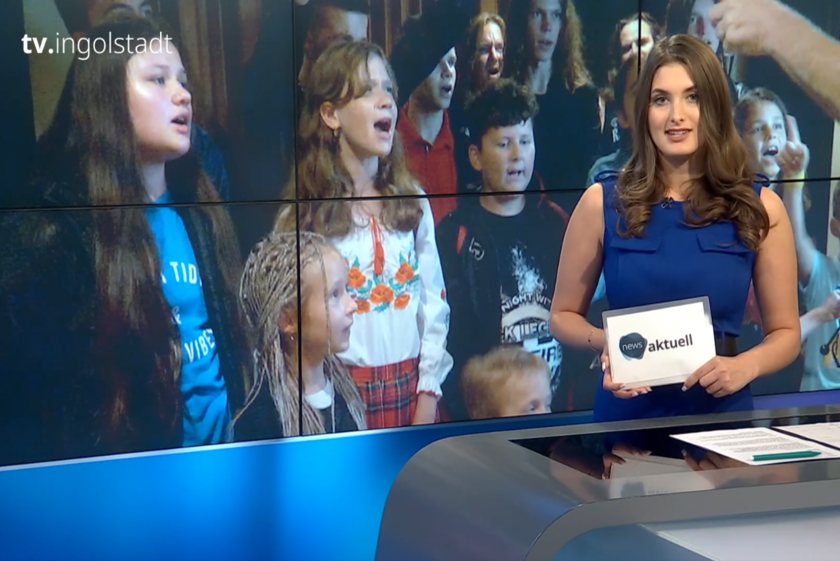 Nachrichtensprecherin im Studio von tv.ingolstadt präsentiert eine Sendung, im Hintergrund ist ein Kinderchor auf dem Bildschirm zu sehen.