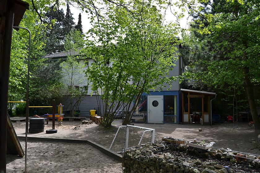 Vor dem Kitagebäude gibt es Sand, Bäume, ein kleines Fußballtor und Hochbeete mit Kräutern.