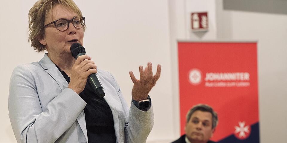 Eine Frau spricht mit einem Mikro in der Hand. im Hintergrund sitzt ein Mann und hört ihr zu.
