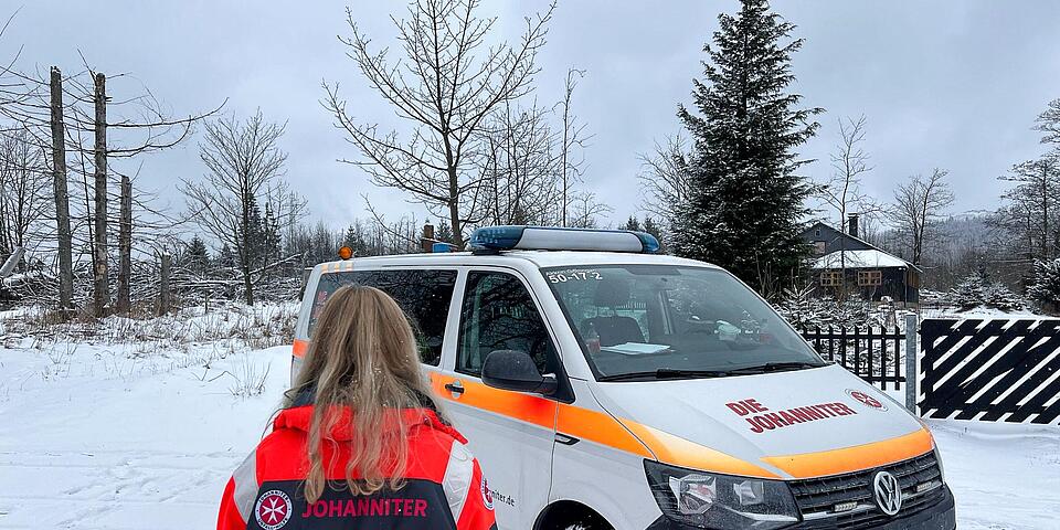 Eine Johanniterin von hinten vor einem Johanniter-Mannschaftstransportwagen in verschneiter Landschaft.