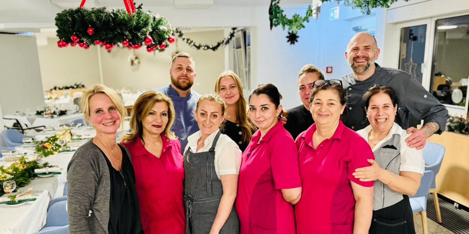 Gruppenbild des Service- und Küchenteams vor festlich geschmücktem Speisesaal, Personal trägt Arbeitskleidung und lächelt in die Kamera.