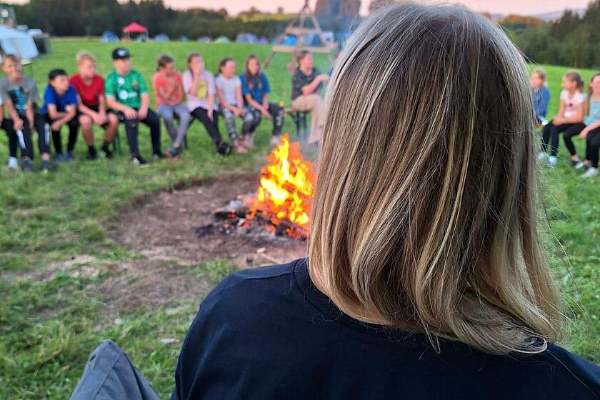 Man sieht eine Frau von hinten, auf ihrem T-Shirt steht „Zeltlagerteam“. Sie sitzt zusammen mit mehreren Kindern in einem Kreis um ein Lagerfeuer.