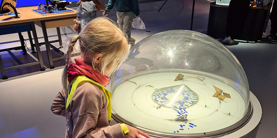 Ein Mädchen schaut auf eine Plexiglashalbkugel auf einer Stele unter der kleine Exponate sind..