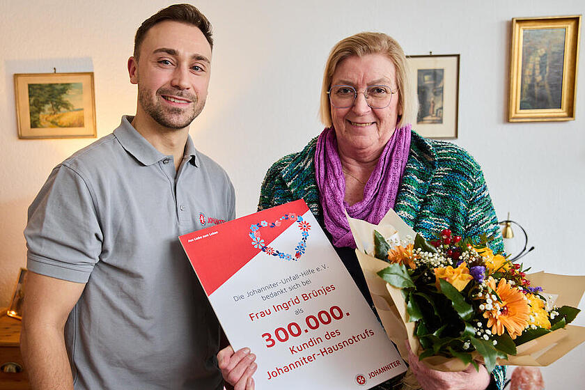 Ein Mitarbeitender der Johanniter überreicht der 300.000 Hausnotruf-Kundin der Johanniter in ihrer Wohnung einen Blumenstrauß und eine Dankesurkunde der Johanniter.
