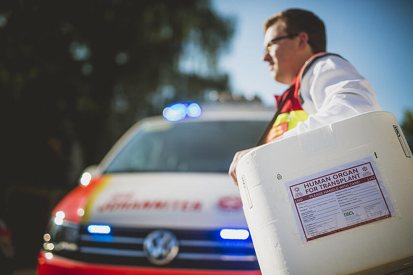 Ein Mann in Dienstkleidung läuft vor einem Johanniter Einsatzfahrzeug entlang und trägt dabei eine Box für Organtransplantationen.