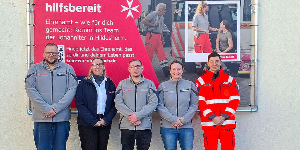 Zwei Männer und eine Frau in grauer Johanniterjacke, eine Frau mit weißer Bluse und blauer Johanniterjacke und ein Mann mit Rettungsdienstbekleidung vor einer Plakatwand auf der steht "Gemeinsam hilfsbereit"
