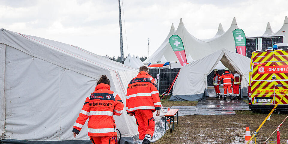 Zwei Johanniter‑Einsatzkräfte in Einsatzkleidung mit der Aufschrift „Bevölkerungsschutz“ gehen zwischen Einsatzzelten über matschigen Boden.