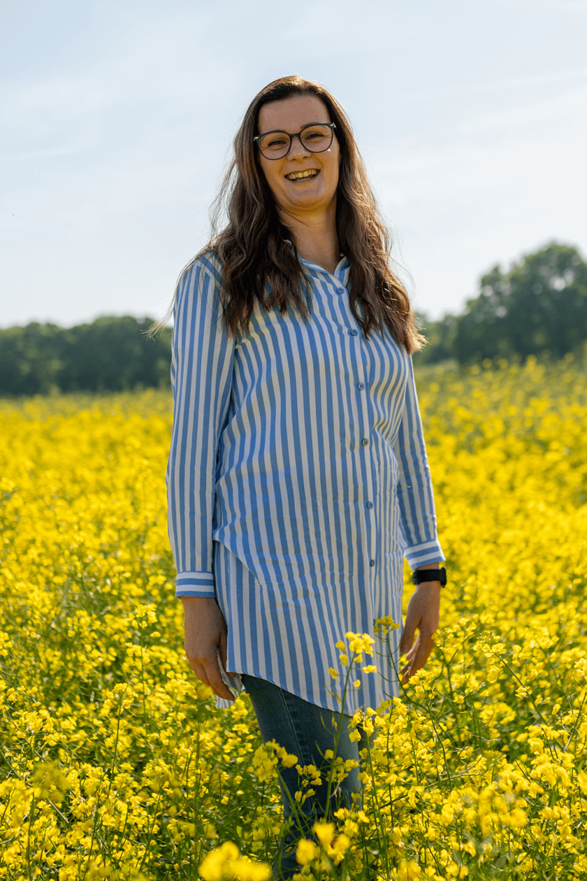 Frau steht in einem Rapsfeld und lächelt, weite gelbe Blumen im Hintergrund.  