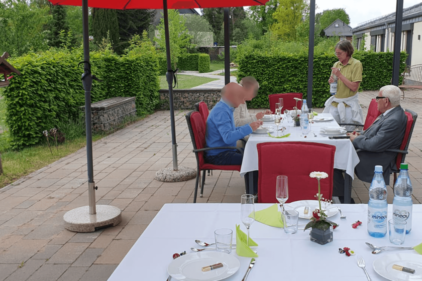 drei Personen sitzen essend unter Sonnenschirmen auf einer Terrasse. Eine vierte Person steht und hält eine Flasche Wasser in den Händen.