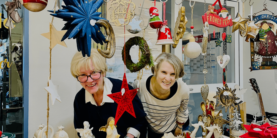 Zwei Frauen stehen hinter einem bunt geschmückten Weihnachtstisch mit vielen handgefertigten Dekoartikeln wie Sternen, Engeln, Kerzen und Weihnachtsfiguren, eingerahmt von hängenden Dekorationen.