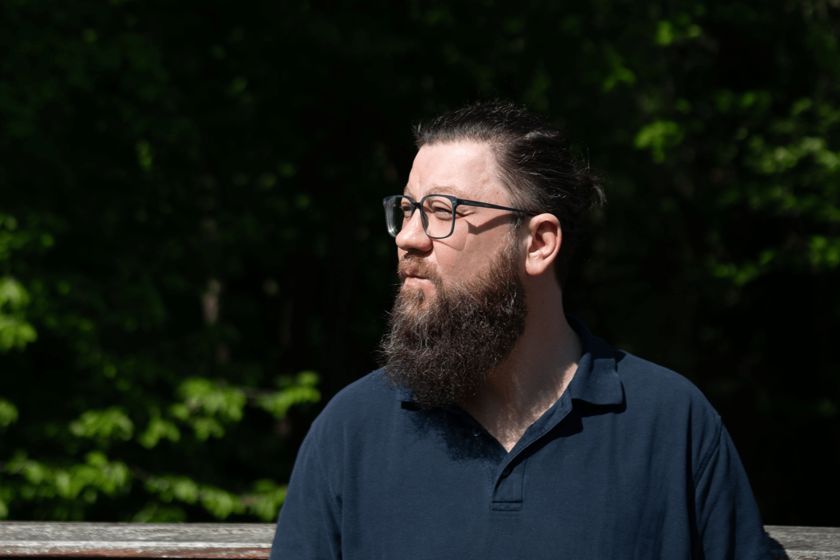Ein bärtiger Mann im dunklen Poloshirt draußen, schaut nach rechts 